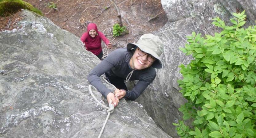 A person who is rock climbing looks up at the camera and smiles. You can also see someone standing on the ground below the climber. 